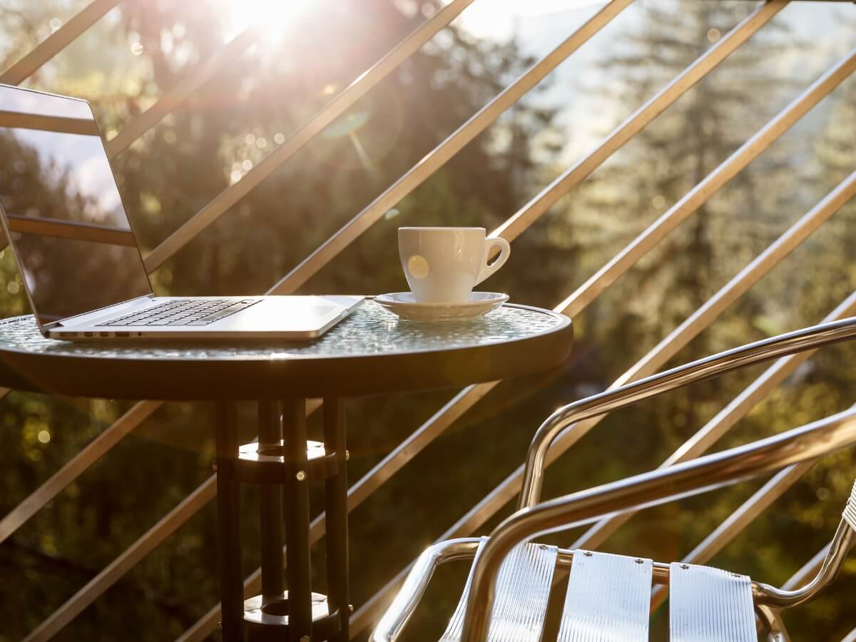 Kovovo-sklenený stolík na slnečnom balkóne s kávou a notebookom.