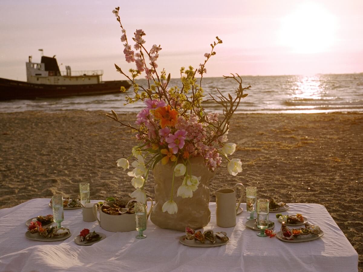 Minimalistické prestieranie s občerstvením na pláži s kvetmi, v pozadí more a loď.