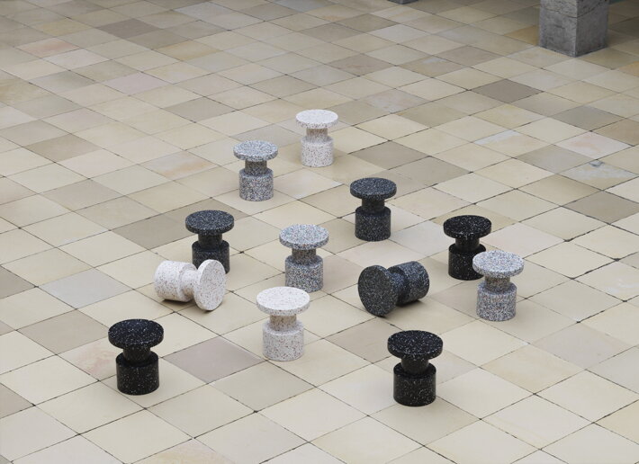 Niekoľko plastových príručných stolíkov/stoličiek rozmiestnených rôzne po štvorcovej dláždenej podlahe